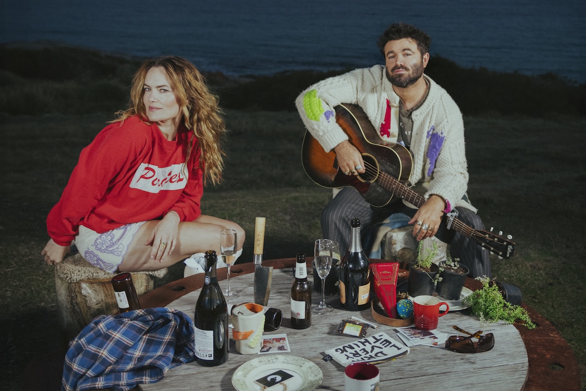 Australian music duo Angus and Julia Stone outdoors at dusk with an acoustic guitar and drinks on a wooden table near the ocean.